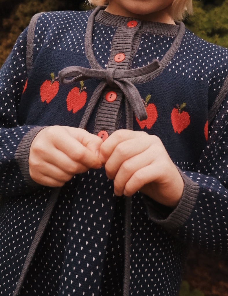 Child wearing a navy sweater with red apple patterns and a gray tie, holding hands together.