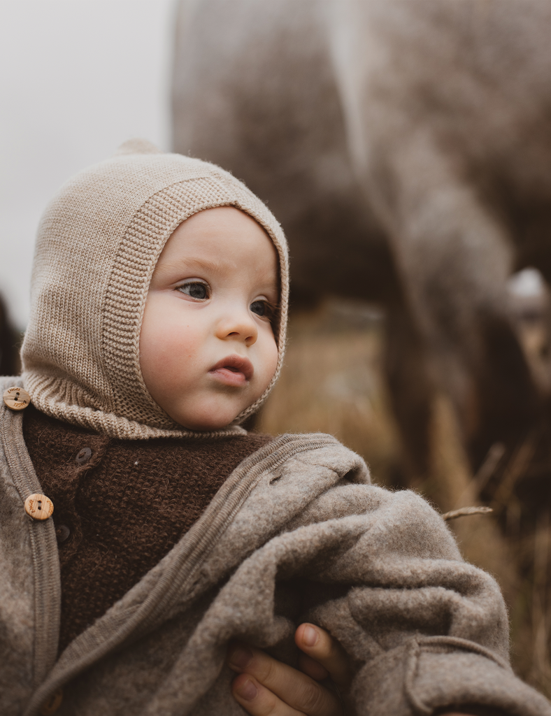 Child wearing a beige knitted hat and coat with a blurred animal in the background