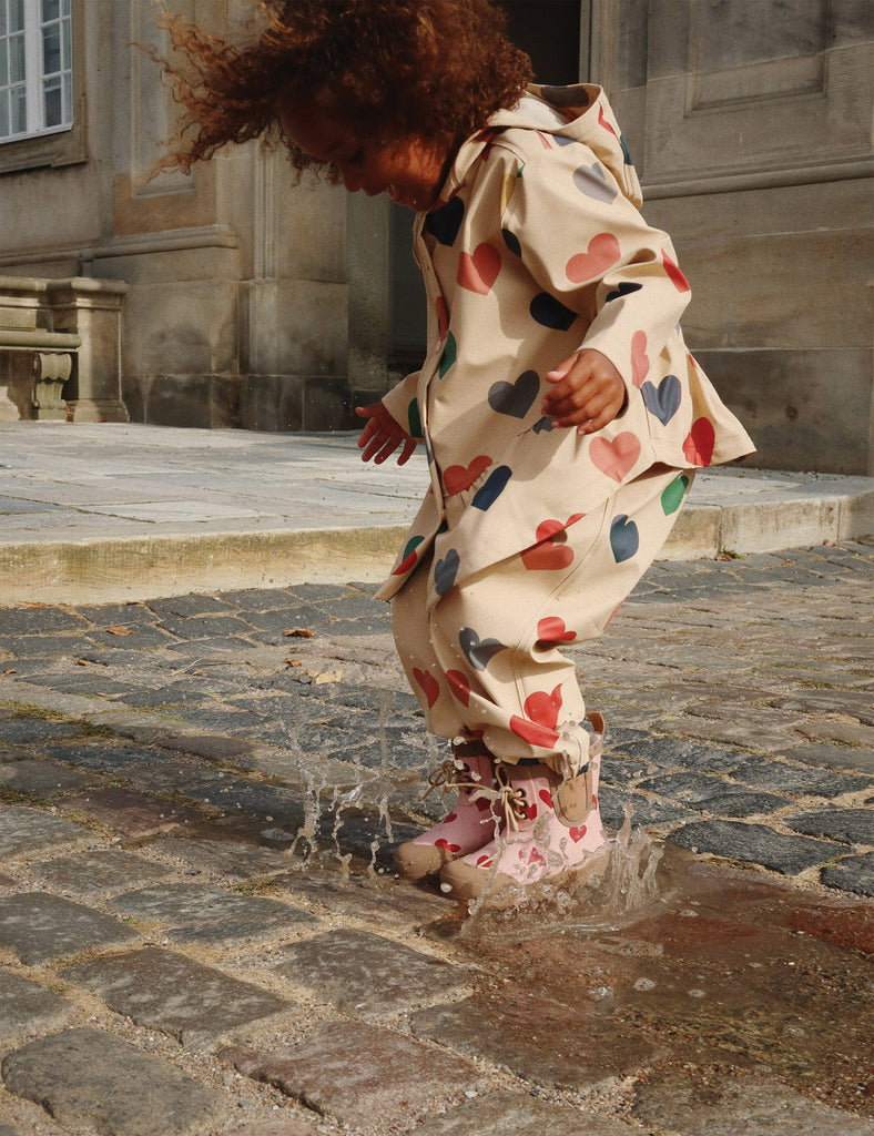 Child in a heart-patterned coat and boots jumping into a puddle on a stone pavement.