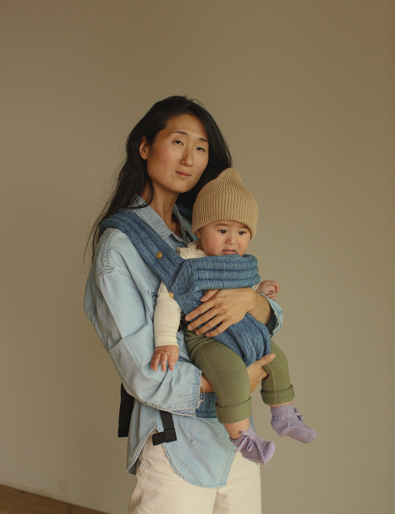 Woman holding a baby in a blue carrier against a beige background