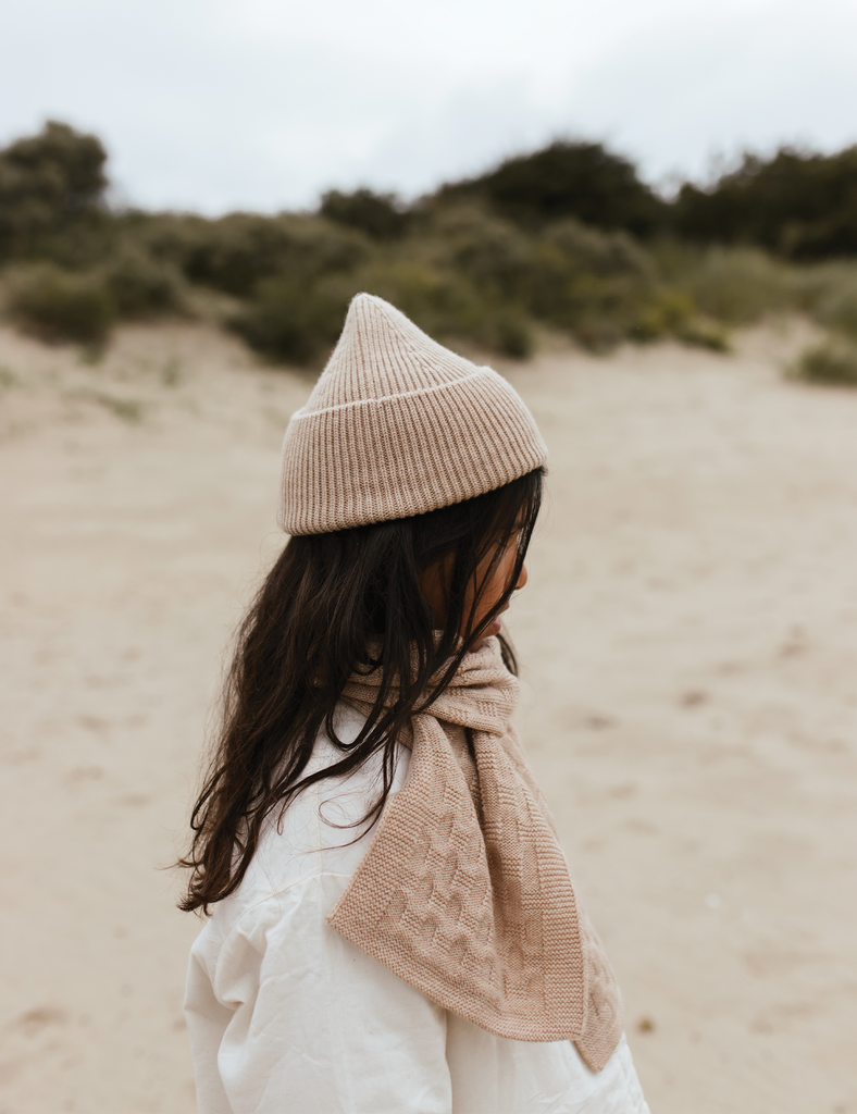 Person wearing a beige knit hat and scarf on a sandy beach with greenery in the background