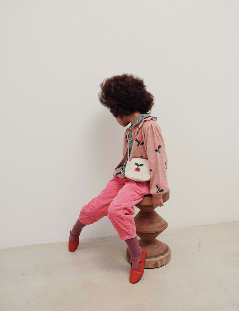Child in pink outfit with red shoes standing on a wooden stool against a white wall.