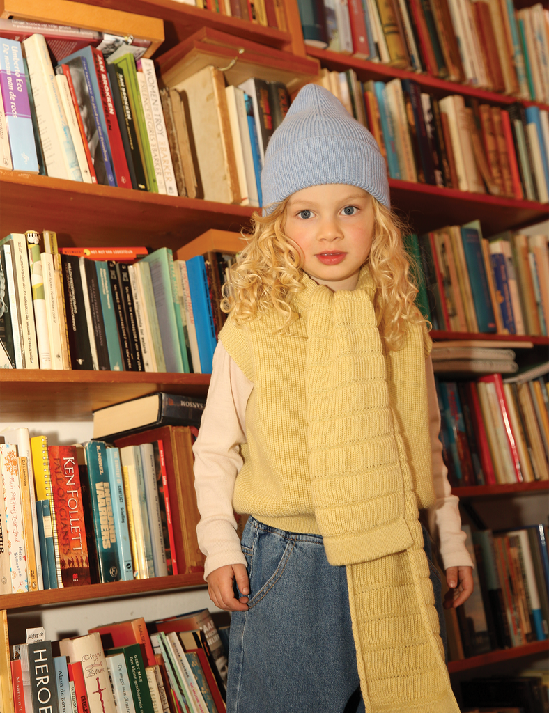 Image of a little girl wearing a blue knitted beanie standing in front of a book shelf.