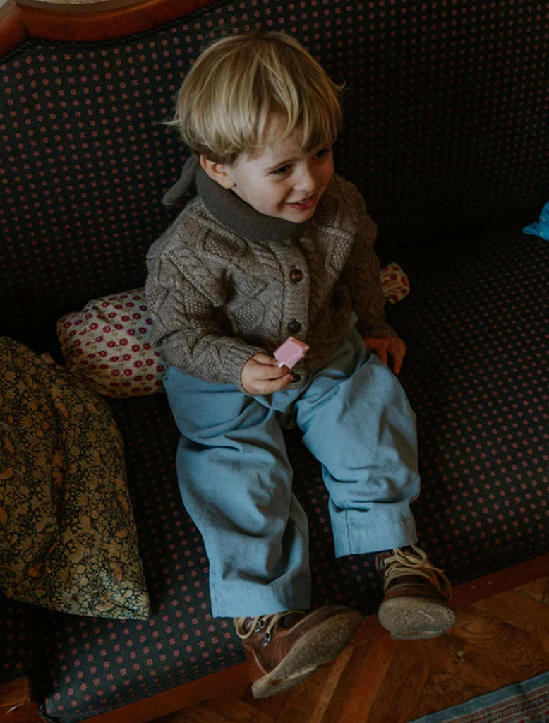 Child in a brown sweater and blue pants sitting on a patterned couch holding a pink object.