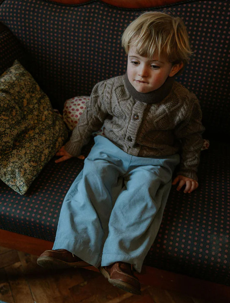 Child sitting on a patterned couch wearing a textured sweater and light blue pants.