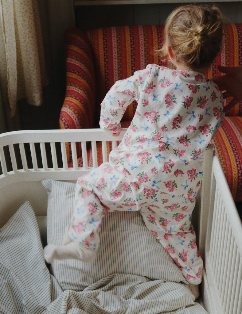 Child in floral pajamas sitting in a crib with a colorful striped chair in the background.