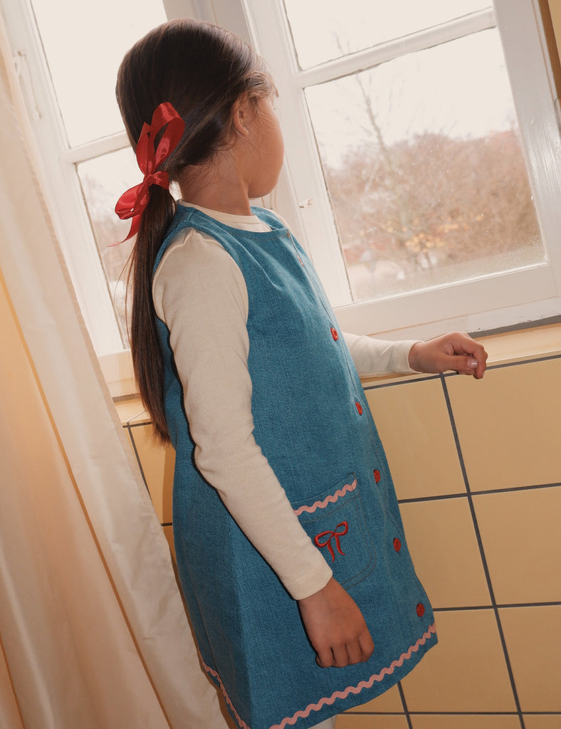 Child wearing a blue dress with red buttons in a room with tiled floor and window.