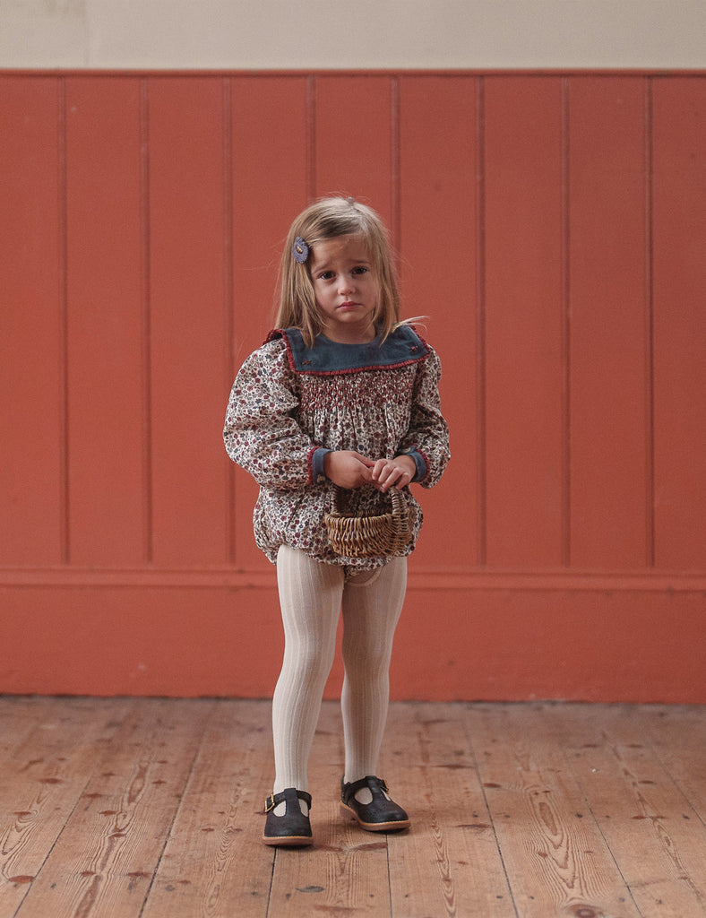 Young girl in a patterned outfit standing against a red paneled wall.