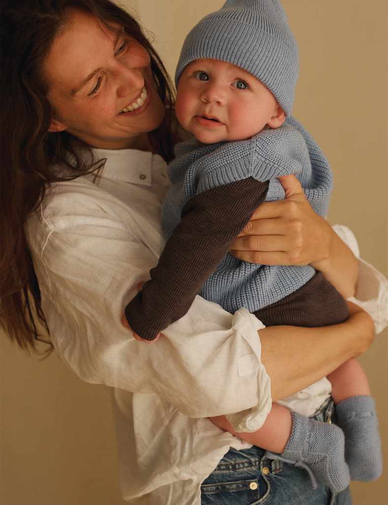 Woman holding a baby wearing a blue knitted outfit against a beige background