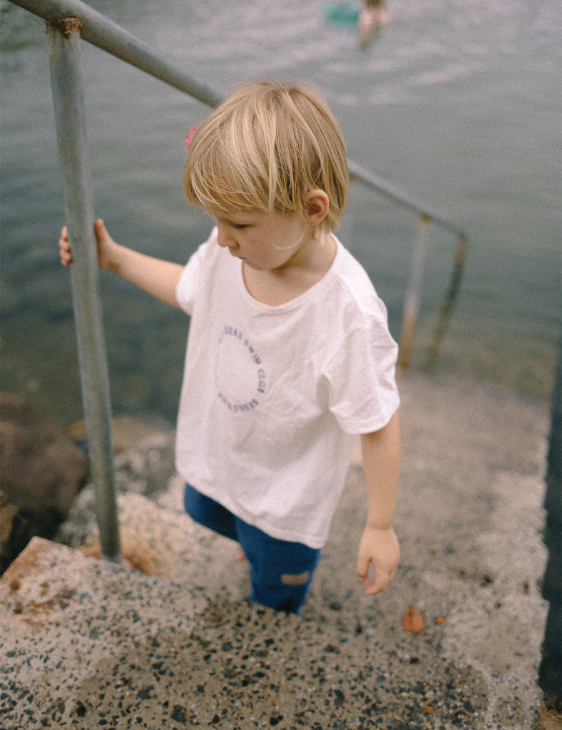 Child in a white shirt and blue pants standing on steps near water