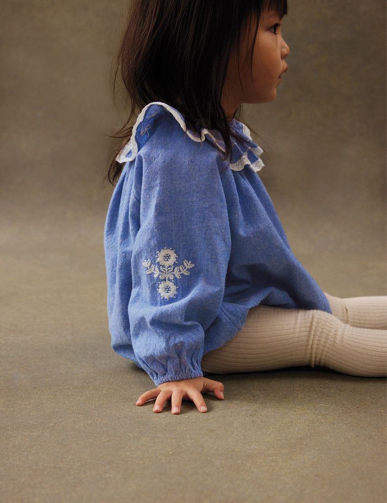 Child wearing a blue embroidered shirt sitting on a beige surface