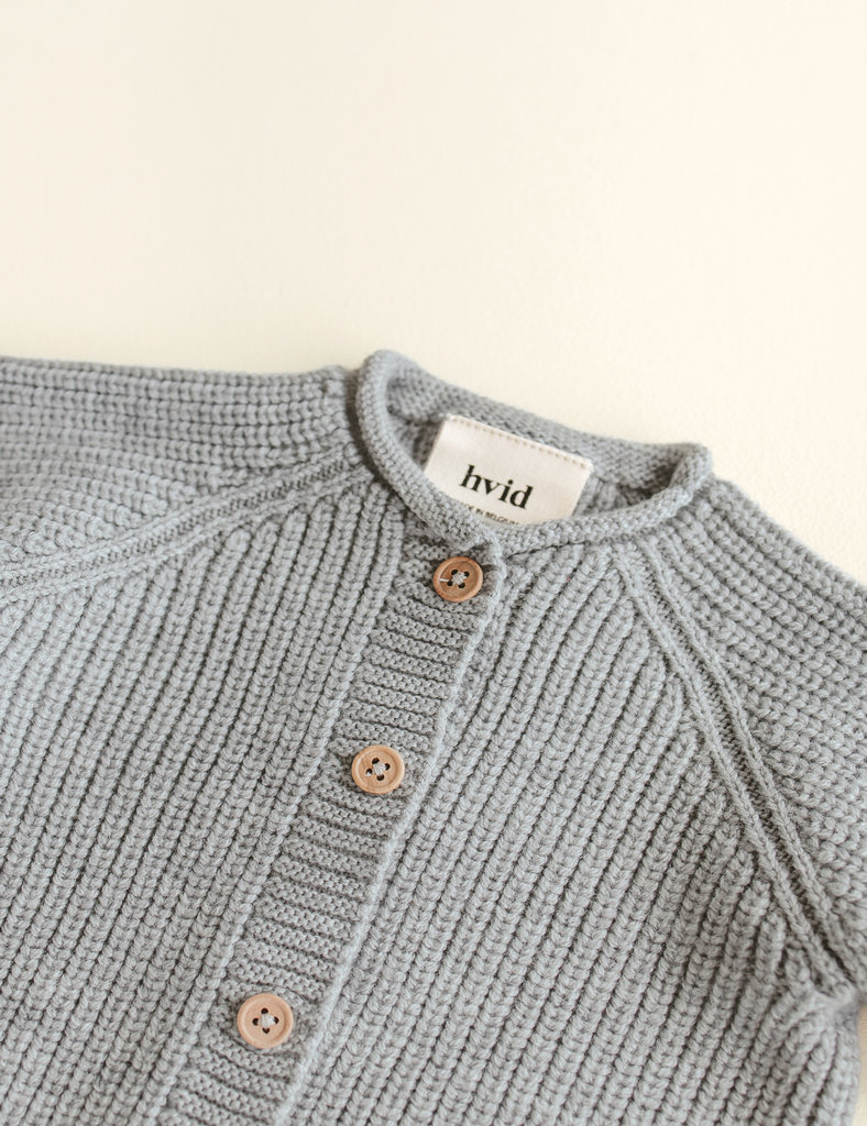 Close-up of a gray knitted cardigan with wooden buttons on a beige background, featuring the brand 'hvid'.