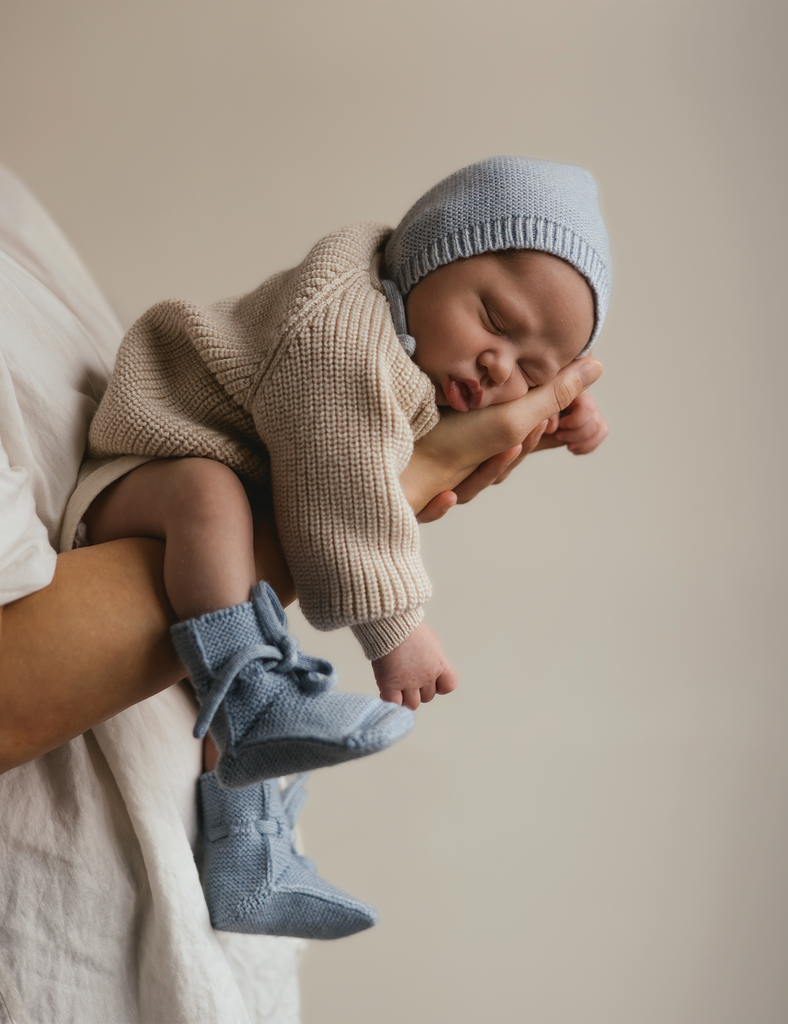 Baby in beige outfit and blue knitted hat and booties being held against a neutral background