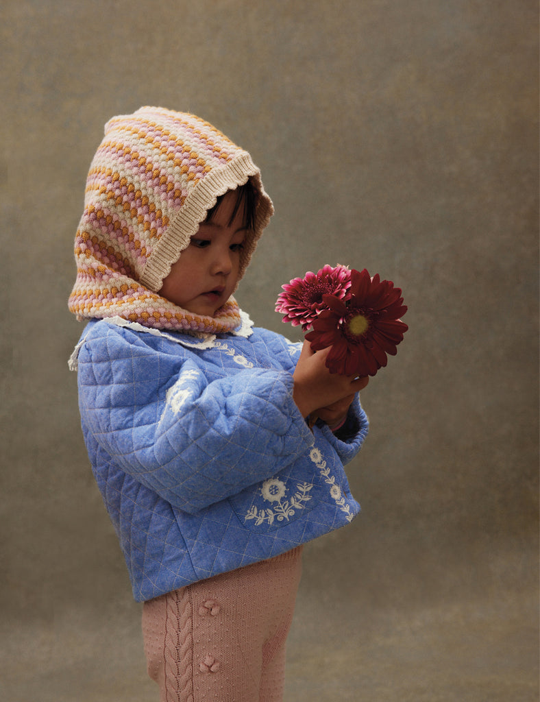 Child wearing a blue quilted jacket and striped hood holding a red flower against a brown background
