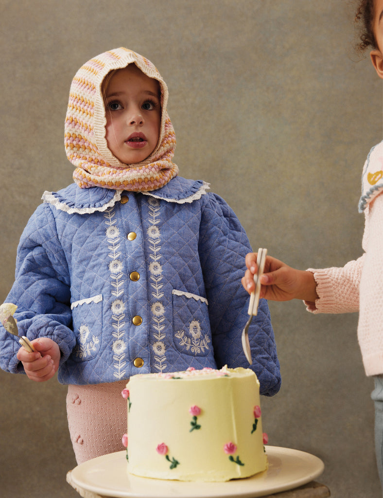 Child in a blue quilted jacket with floral patterns, standing next to a small cake on a plate.