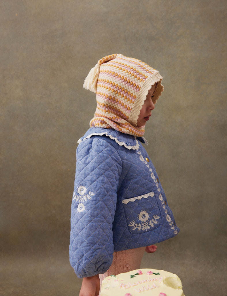 Child wearing a blue quilted jacket with floral embroidery and a striped bonnet against a brown background