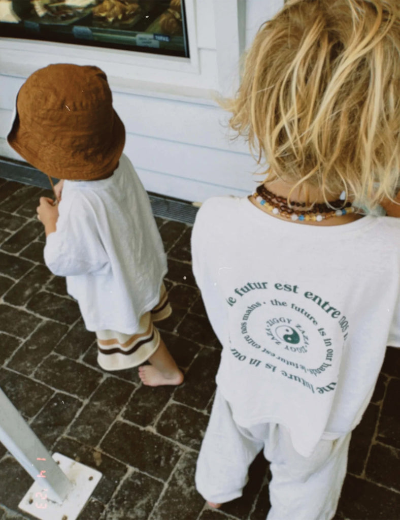 Two children standing on a stone pavement, one wearing a brown hat and white outfit, the other in a white shirt with text.