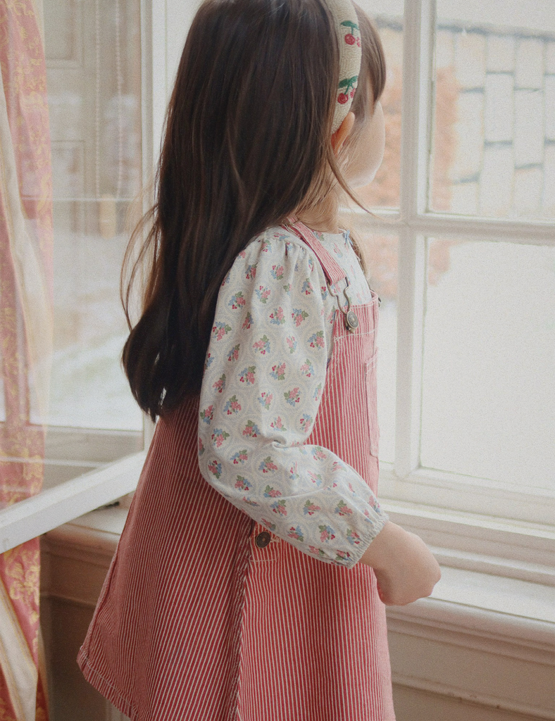 Young girl in a pink dress and floral headband standing by a window.