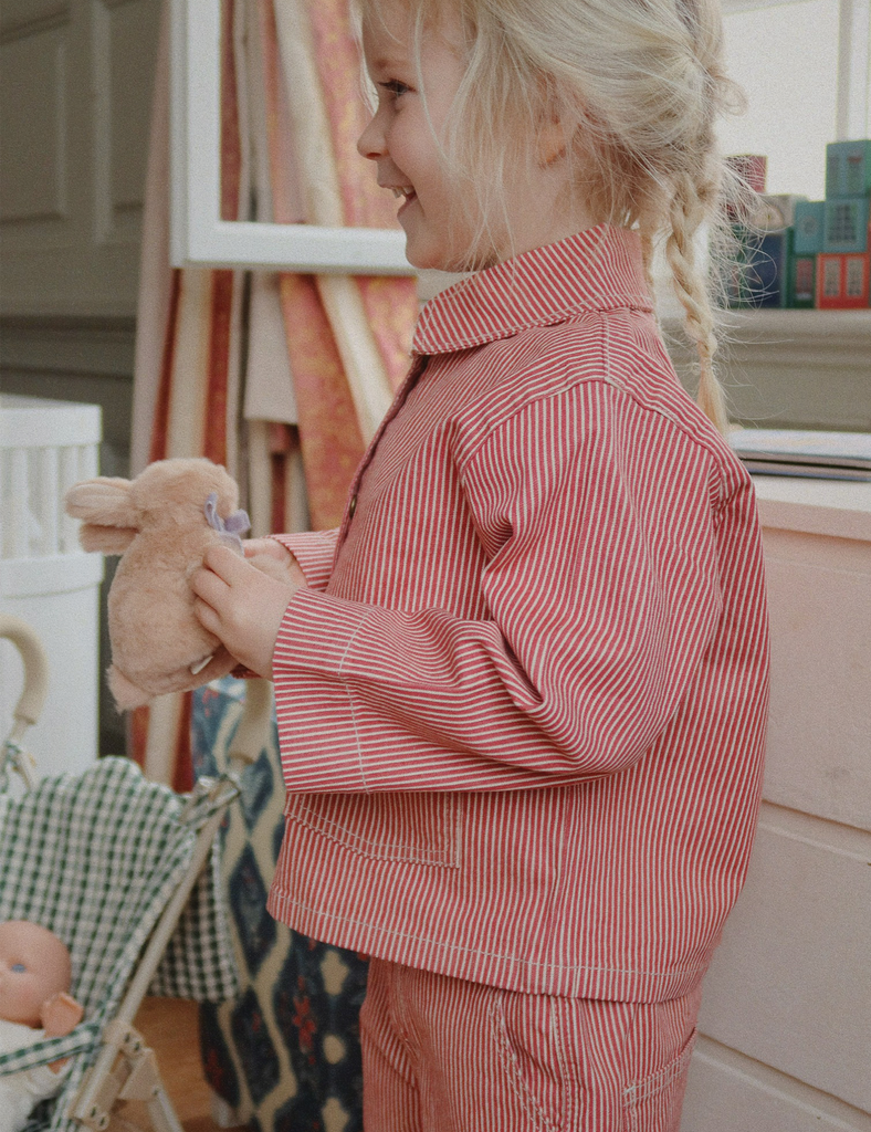 Child in red and white striped pajamas holding a teddy bear in a room with toys and furniture.