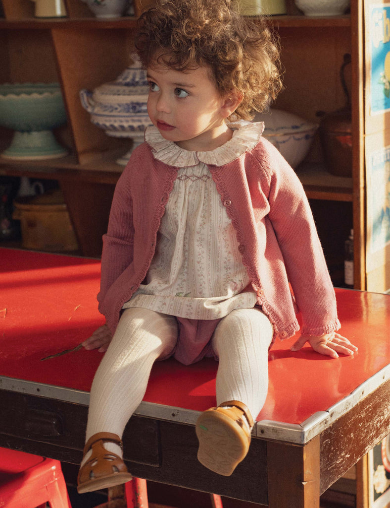 Child sitting on a red table wearing a pink cardigan and white dress.