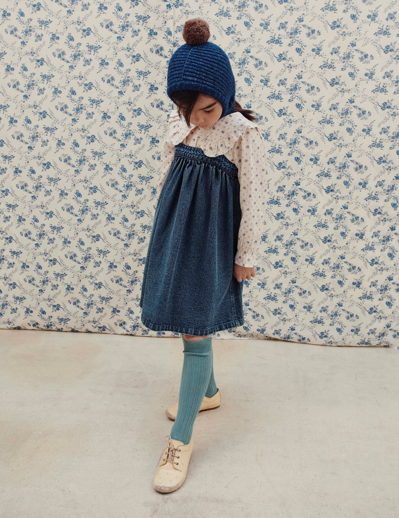 Child wearing a blue dress and hat against a floral wallpaper background