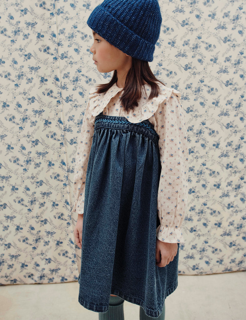 Child wearing a blue knitted hat and denim dress against a floral wallpaper background