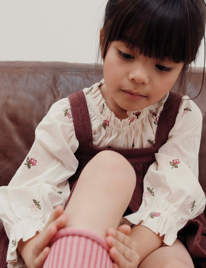 Child wearing a white blouse with floral embroidery and a brown dress sitting on a couch.