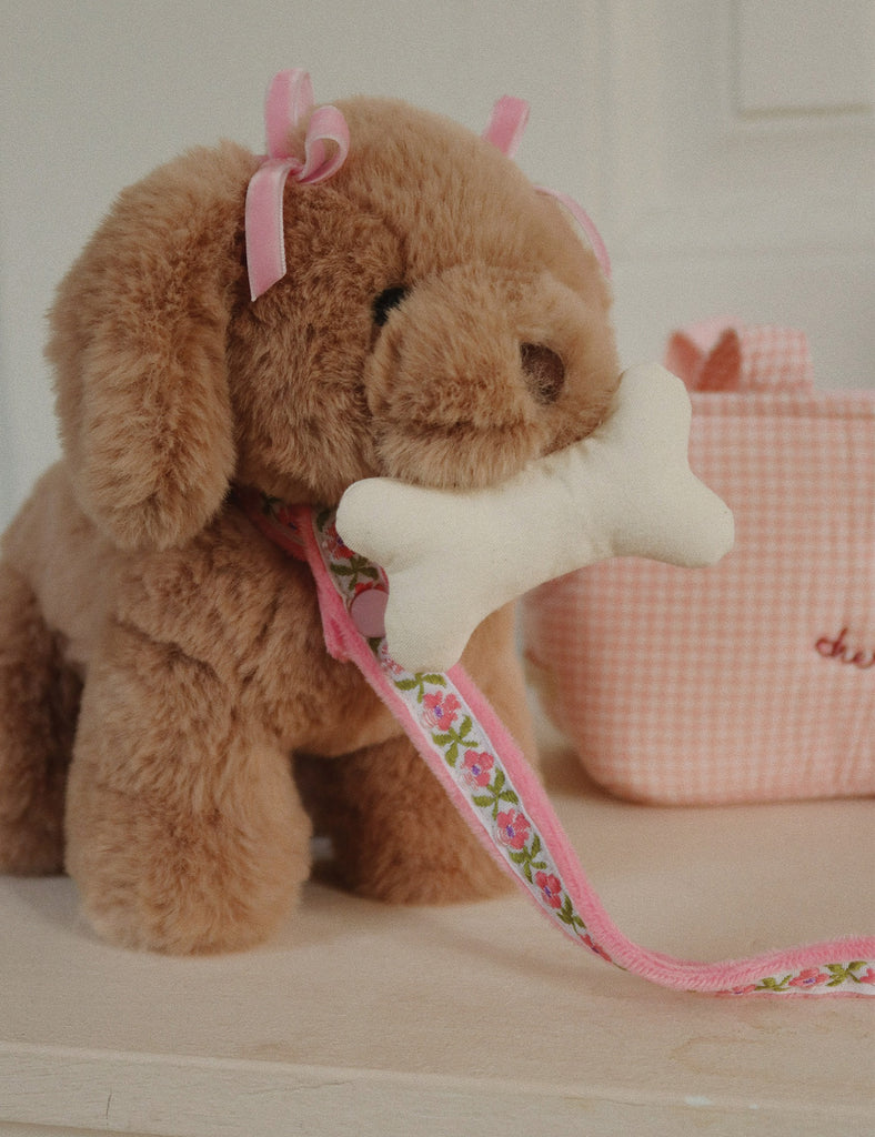 Plush dog with a bone-shaped chew toy on a pink leash, sitting on a light wooden surface.