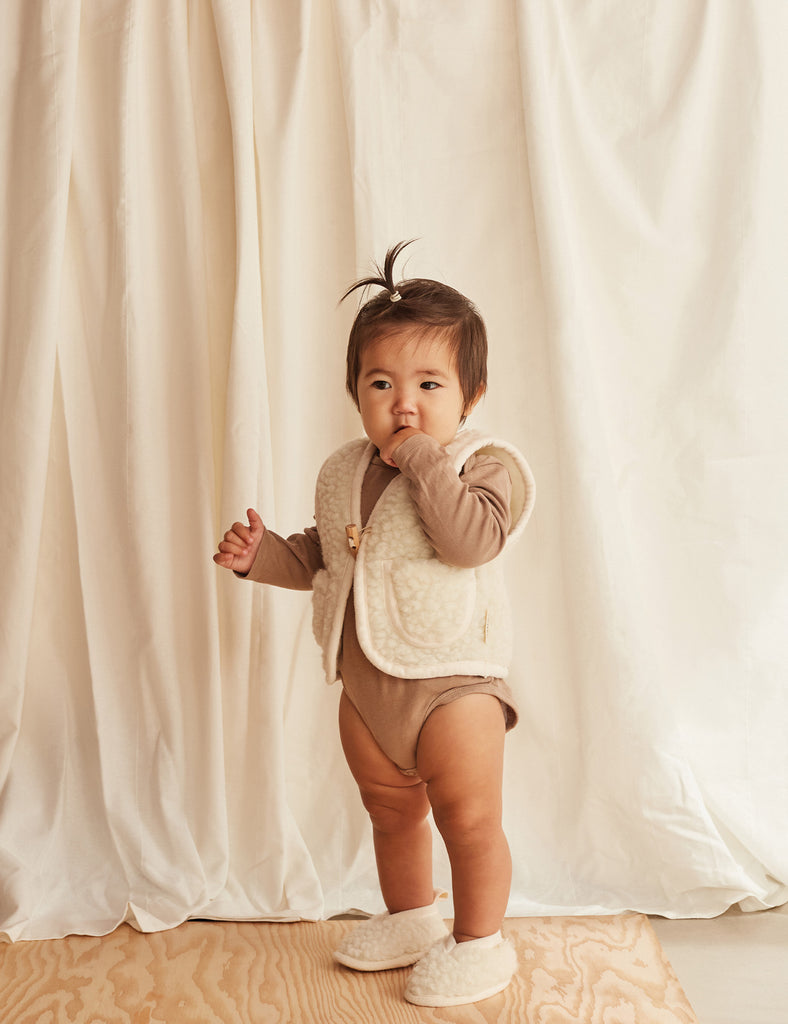 Baby in a brown outfit standing against a white curtain