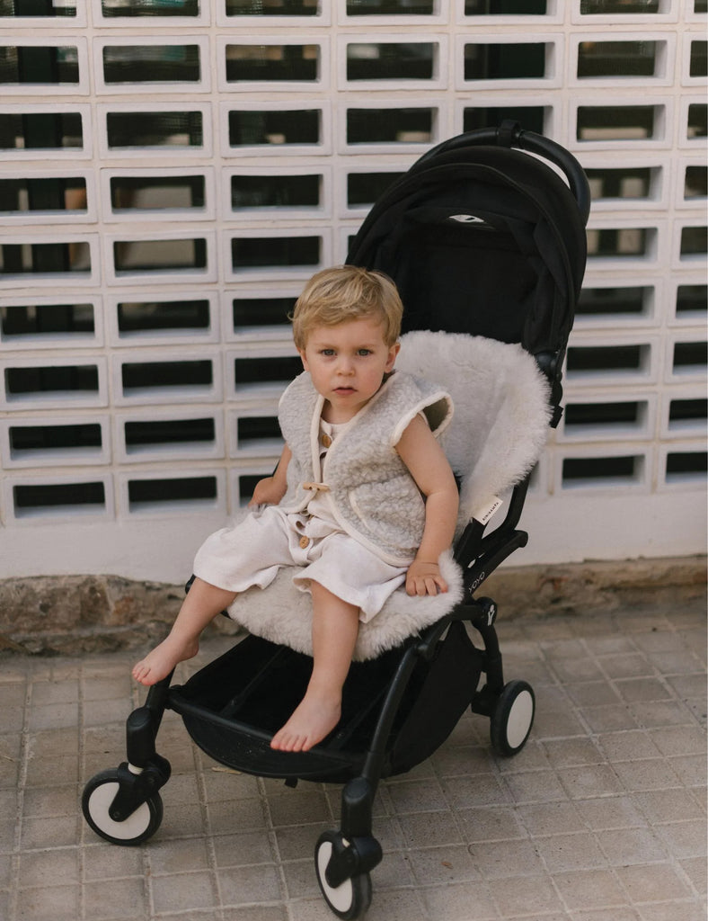 Child sitting in a stroller with a textured stroller cover against a white brick wall.