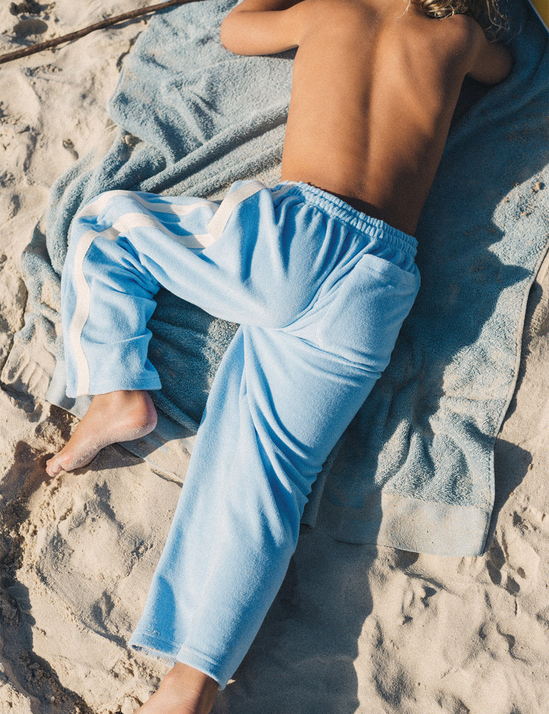 Person wearing light blue sweatpants lying on a towel on a sandy beach.