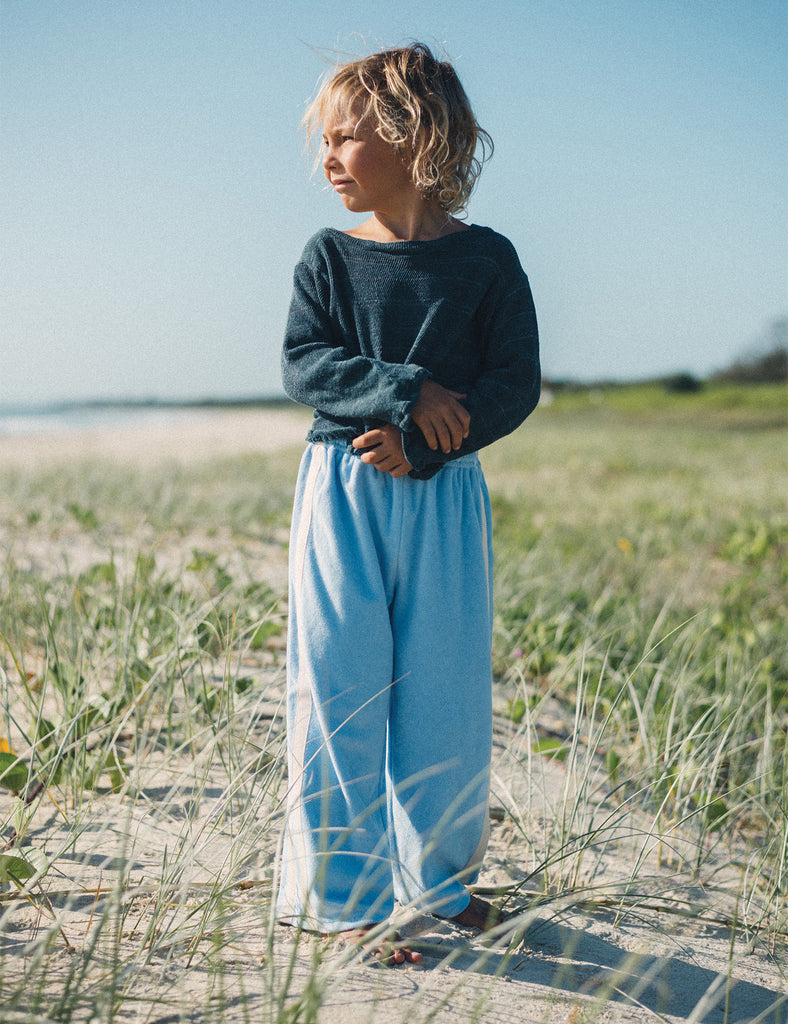 Child standing on a sandy beach wearing a dark sweater and light blue pants.