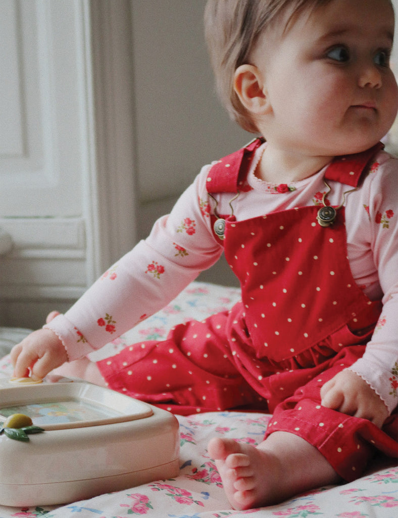 Baby in red polka dot overalls playing with a toy on a floral blanket.