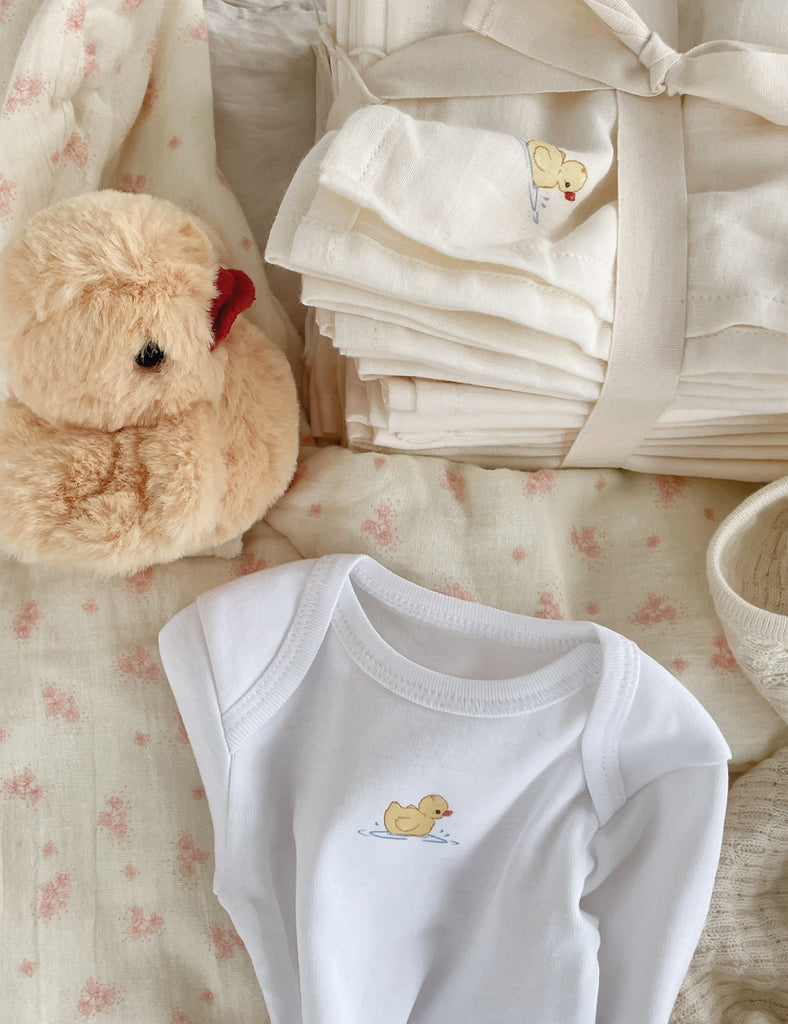 White baby onesie with a duck design, teddy bear, and folded white fabric on a floral blanket.