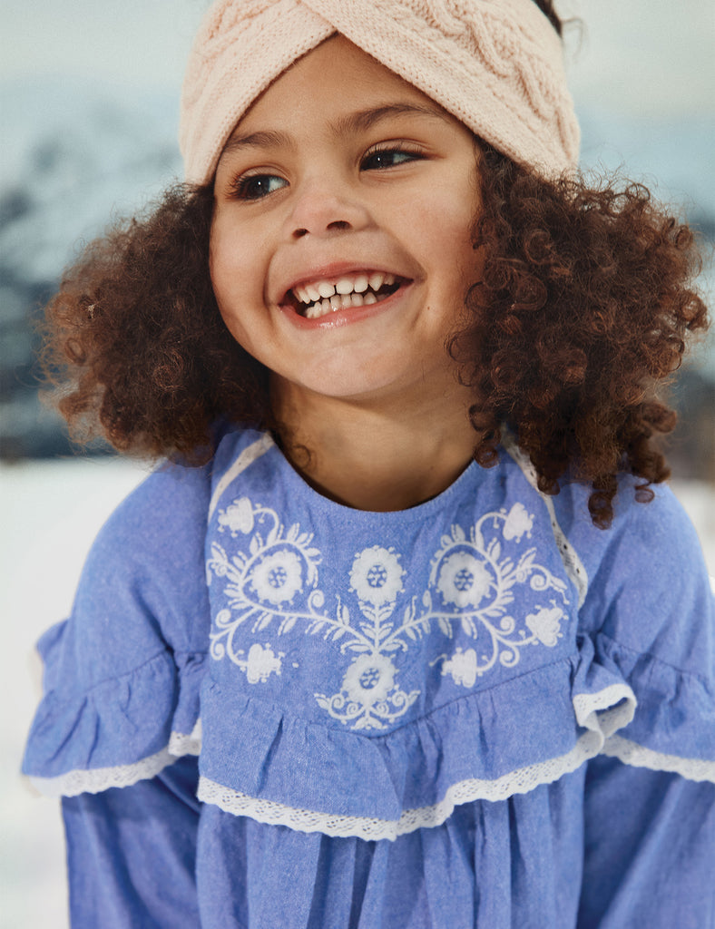 Child wearing a blue dress with white embroidery and a pink headband, smiling outdoors.