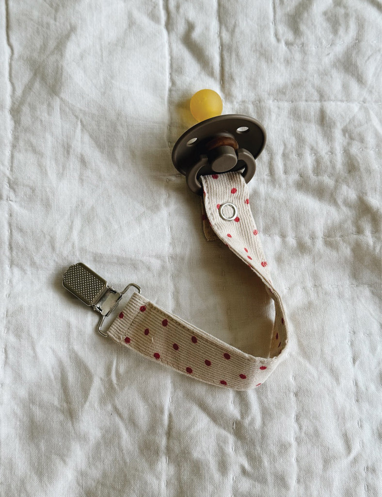 Image of a red dot pacifier clip connected to a brown pacifier on  cream background.