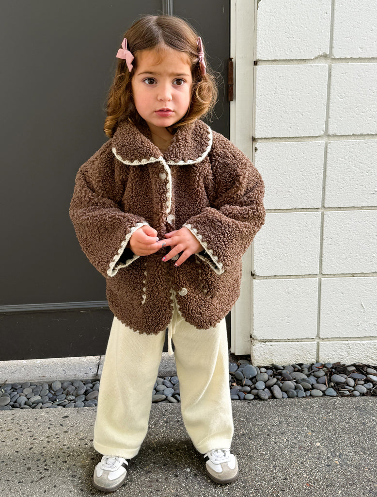 Sloane wears the Paddy Jacket in Brown in size 2Y. She is wearing cream colored knit pants, sneakers and two pink bows in her hair.