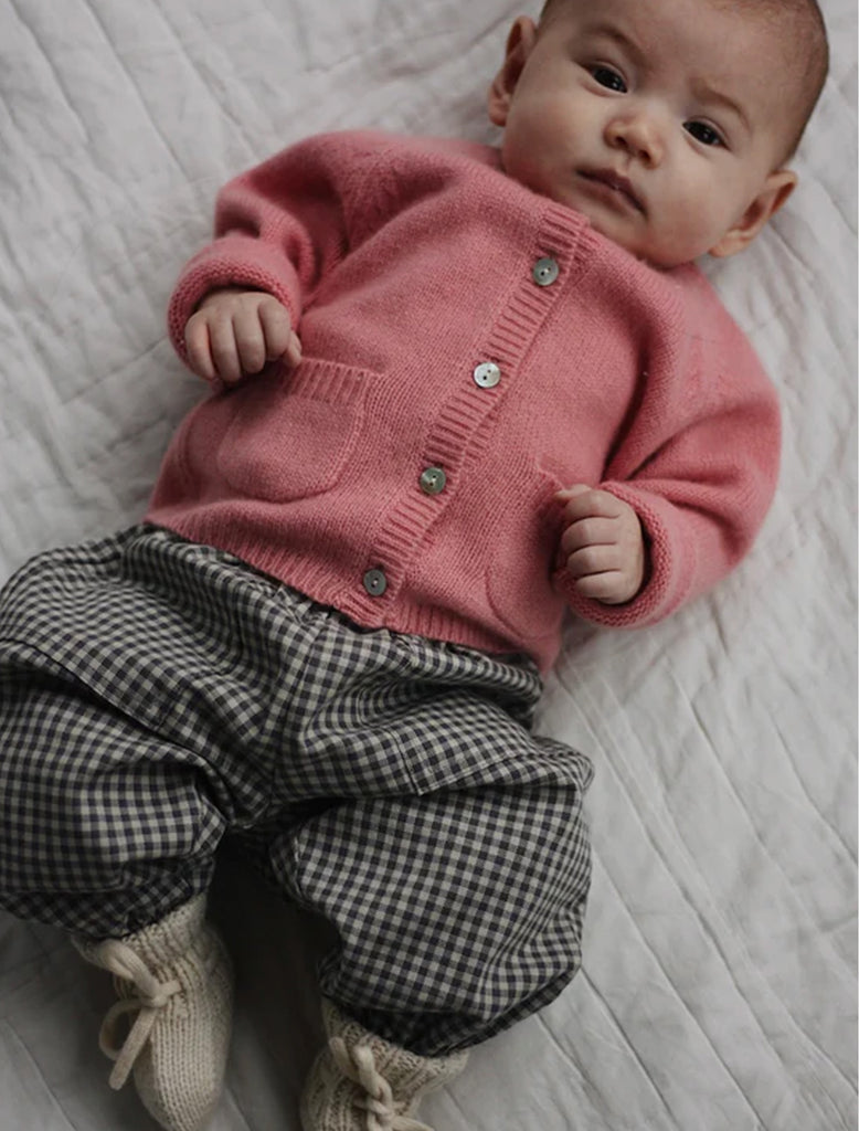 Baby wearing a pink knitted cardigan and checkered pants on a white blanket