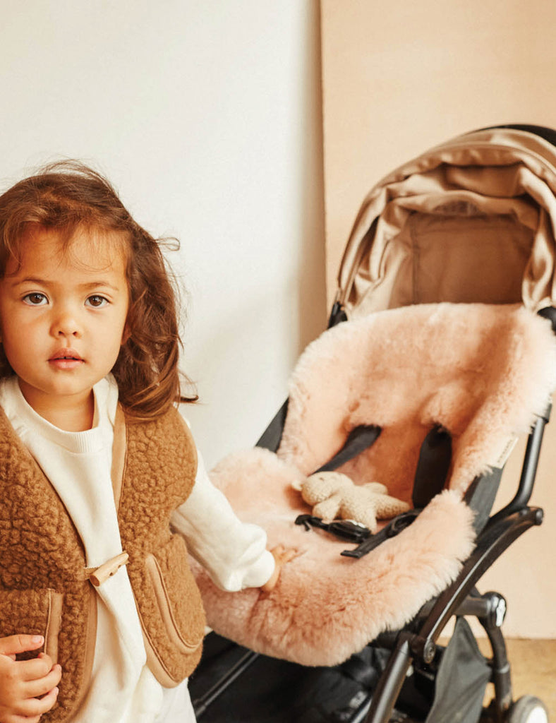 Child in a brown coat standing next to an open beige stroller with fluffy interior.