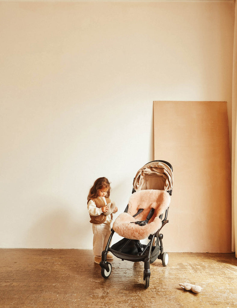 Child standing next to a baby stroller in a minimalistic room with a wooden floor and white walls.