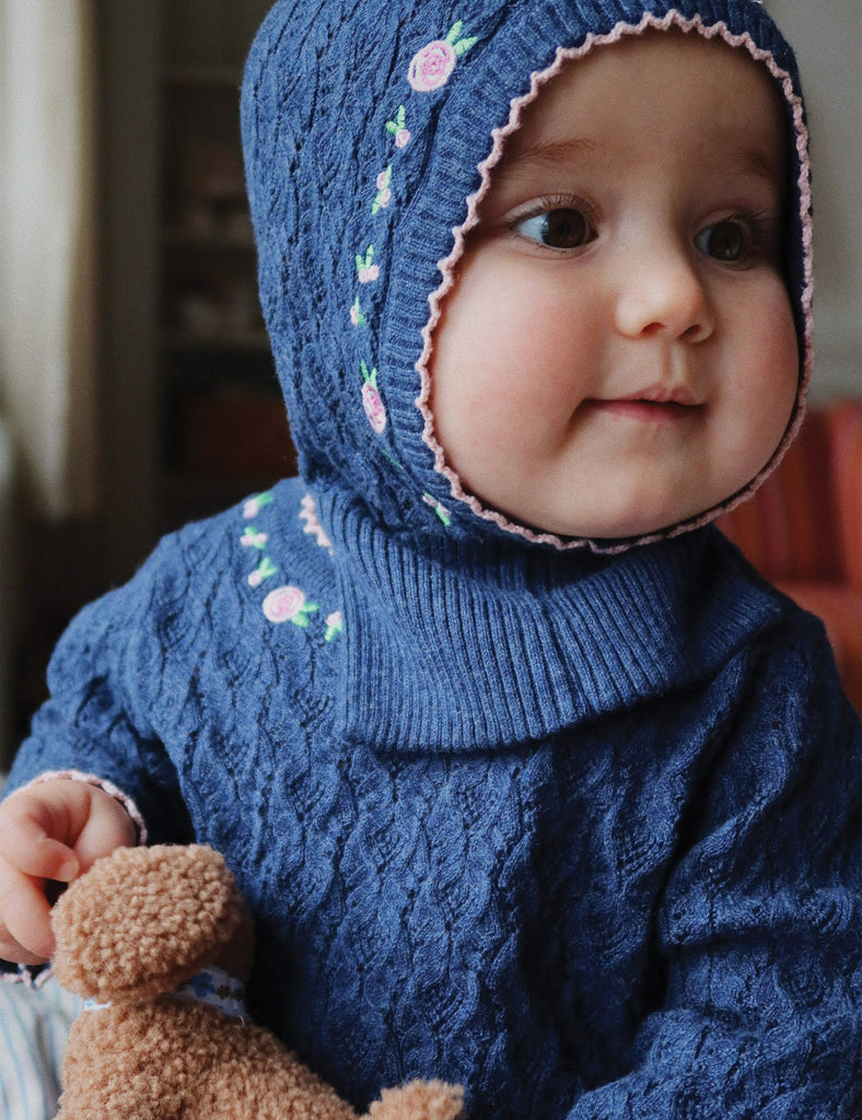 Child wearing a blue knitted outfit with floral patterns, holding a teddy bear.