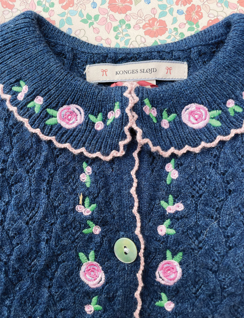 Blue knitted cardigan with floral embroidery and a brand tag on a floral background