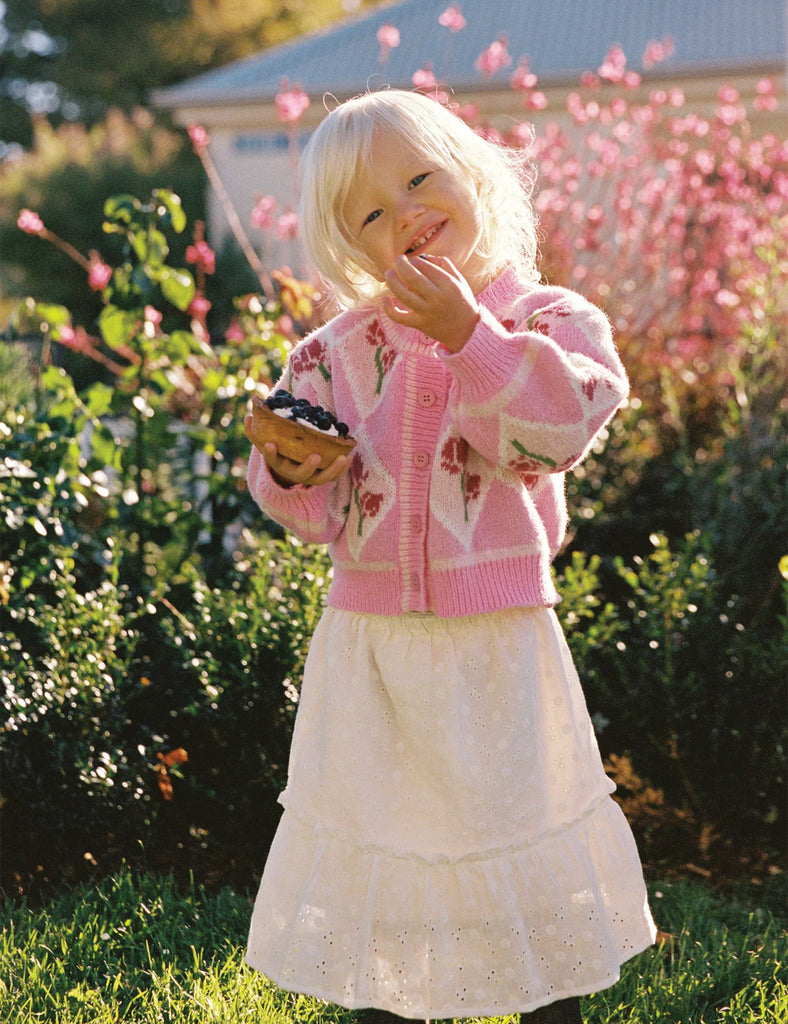Image of a little girl wearing a pink cardigan and white long skirt standing in front of some flowers.