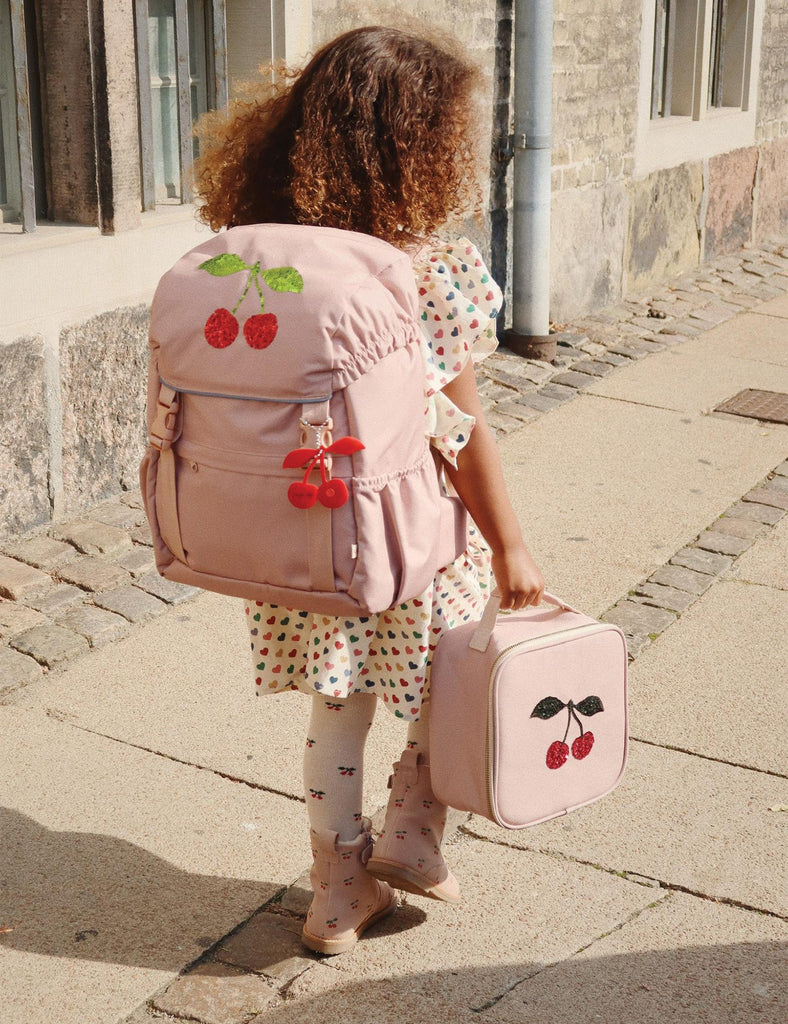 Child with pink backpack and suitcase featuring cherry designs on a stone pavement.