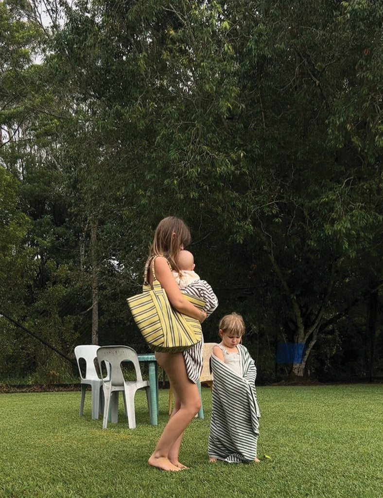 Woman holding a baby and standing next to a child wrapped in a blanket on grass with trees in the background