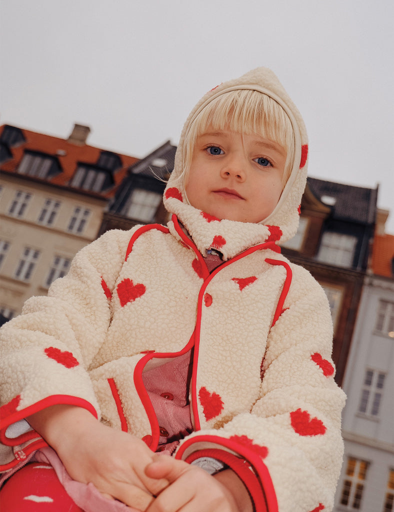 Child sitting down wearing a white fleece jacket with a red heart print on it.