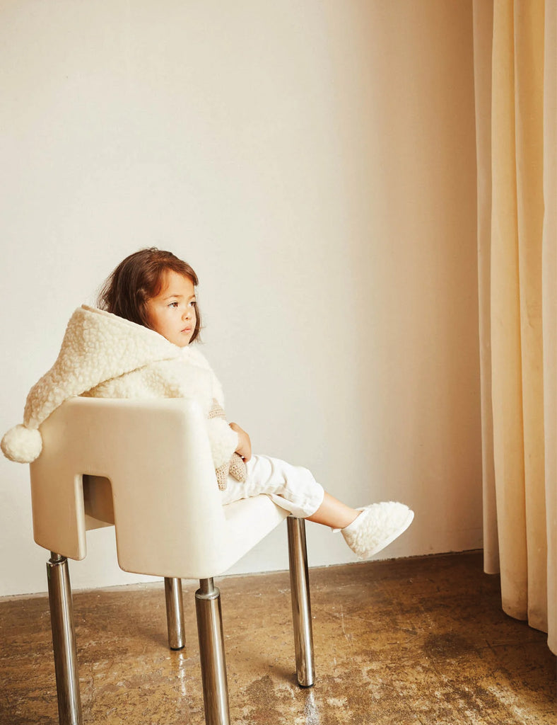 Child sitting on a chair wearing a white coat with a hood in a room with beige walls and curtains.