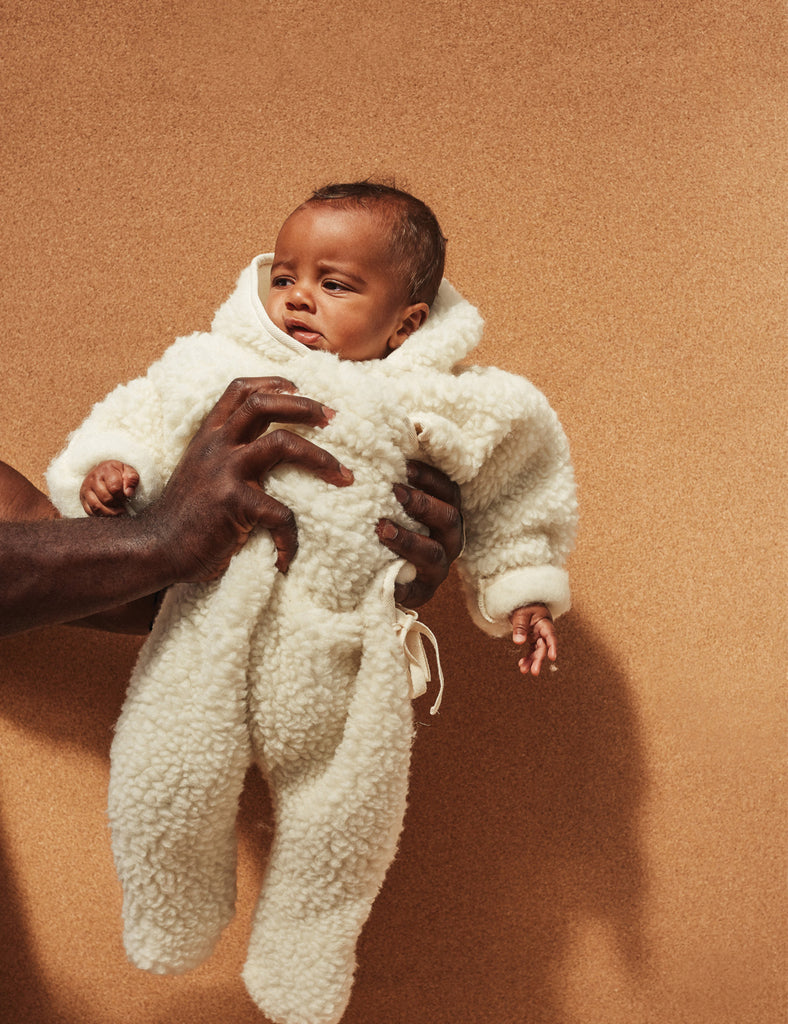 Baby in a white fleece outfit being held against a brown background