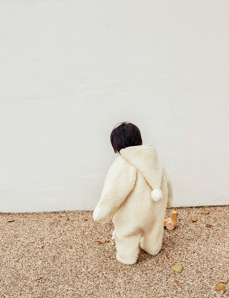 Person wearing a fluffy white coat with a hood on a beige floor against a white wall.