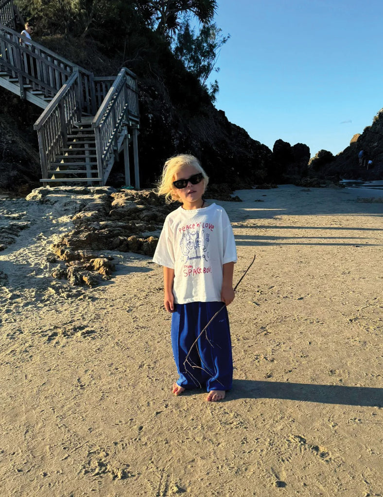 Child standing on a sandy beach holding a stick, with a staircase and rocky landscape in the background.